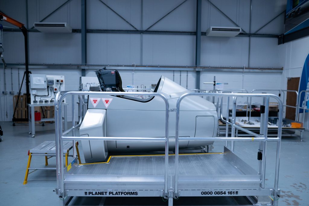 The image depicts an industrial setting, likely a warehouse or manufacturing facility. In the center, there is a large gray industrial machine equipped with safety features such as railings and caution symbols. The machine’s control panel includes an electronic screen and possibly some buttons or switches. It rests on a metal platform labeled “PLANET PLATFORMS.” The background shows the interior of the warehouse with high ceilings, steel beams, and overhead lights. No people are visible in the image.