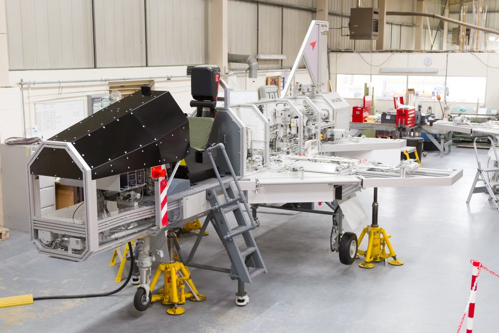 A large metal training piece of equipment. The equipment replicas an airframe. There equipment is in a warehouse workshop with a number of pieces of paper and people working around it.