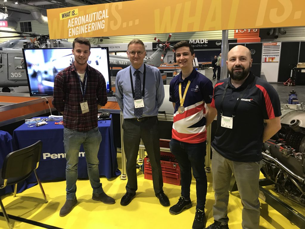 Four people at an exhibition event. They are wearing a mixture of office suite and professional polo shirts. They are stood by a sign that says "What is....Aeronautical Engineering"