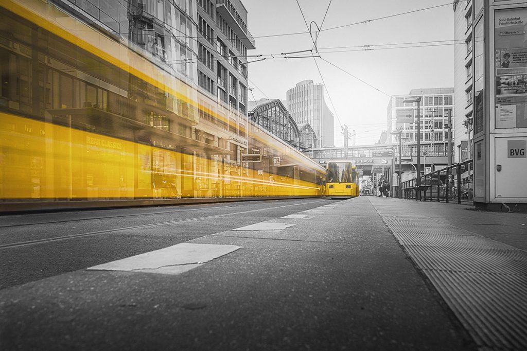 A black and white image of a city street with a tram station, featuring a yellow tram in motion, creating a blur effect as it moves through the scene. The tram lines overhead and multiple tracks on the ground suggest an urban setting. A tram stop sign with the letters “BVG” is visible on the right side. The textured tiles on the ground indicate a tram station platform.