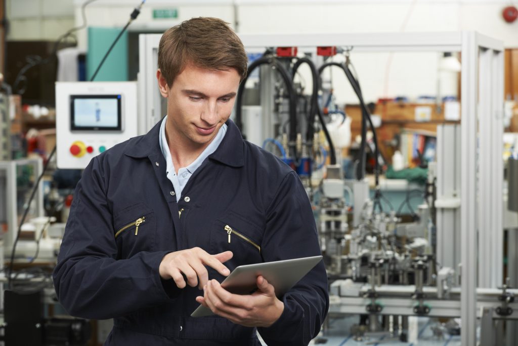 An individual in a dark blue work uniform holds a tablet in an industrial setting with machinery in the background. The person’s face is blurred for privacy. The uniform features visible zippers, and the machinery suggests a factory or manufacturing plant environment. Various tubes, wires, and metal structures are part of the complex assembly or production line.