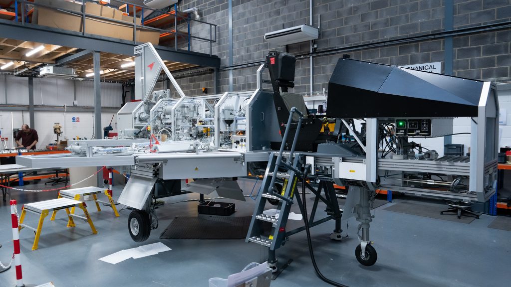 A large metal training piece of equipment. The equipment replicas an airframe. There equipment is in a warehouse workshop with a number of pieces of paper and people working around it.