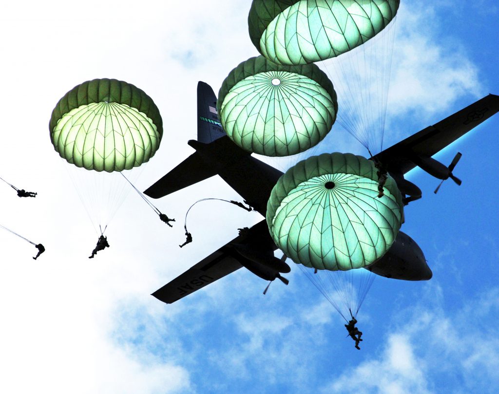 An image of blue sky and clouds. There are four green parachutes in the sky and an aeroplane