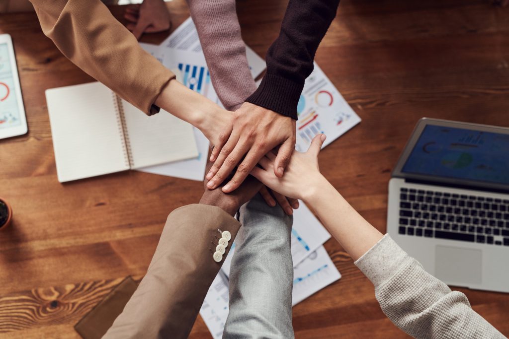 The image depicts a group of people stacking their hands in the center, suggesting unity or teamwork. The hands are of different skin tones, implying diversity. There are business-related papers with charts and graphs scattered on the wooden table around their hands. A laptop is also visible on the table, indicating a work or meeting environment. The sleeves of the individuals suggest a professional or semi-formal attire.