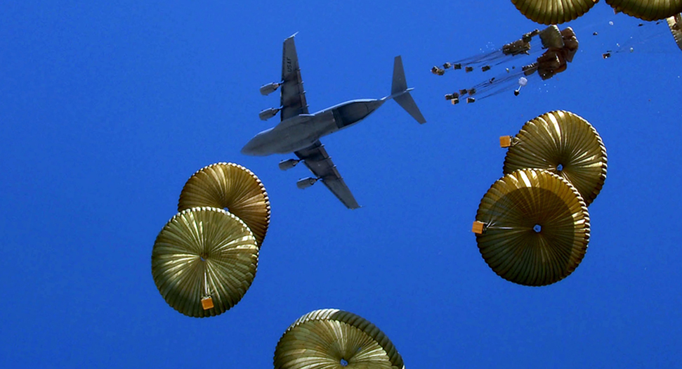 A stock image of a plane dropping numerous parachute parcels. The sky is blue.