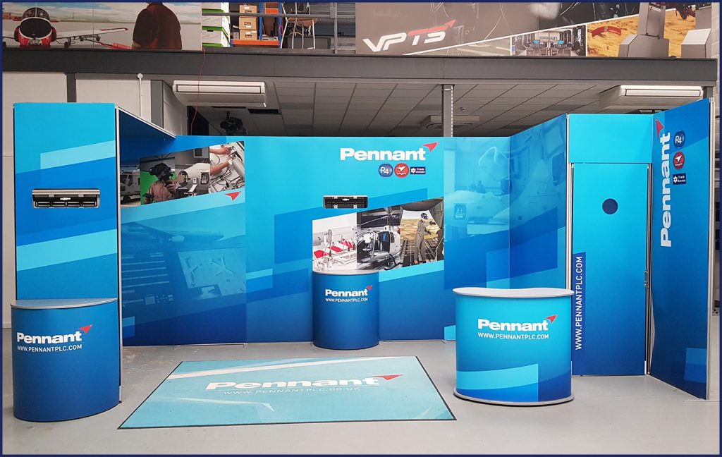 A mock-up design of an exhibitor stand in a large warehouse room. The stand in a multitude of blue colours with the word "PENNANT" on. There are various images on the panels of the stand.