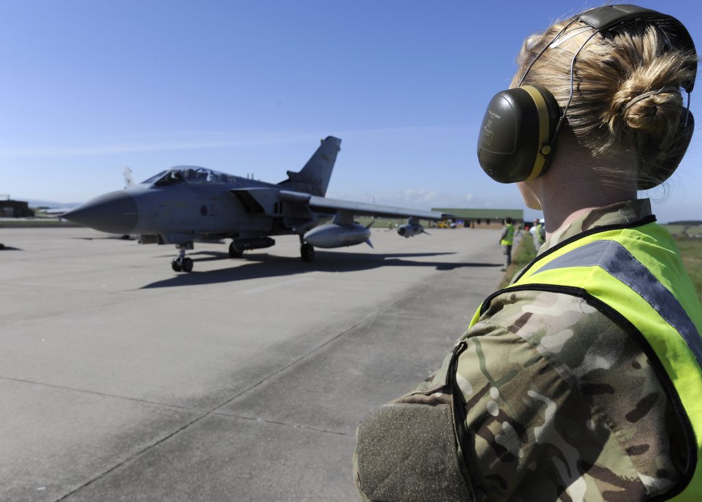 A military airfield with a person in camouflage starring at a jet that is about to take off. It's a day with blue sky.