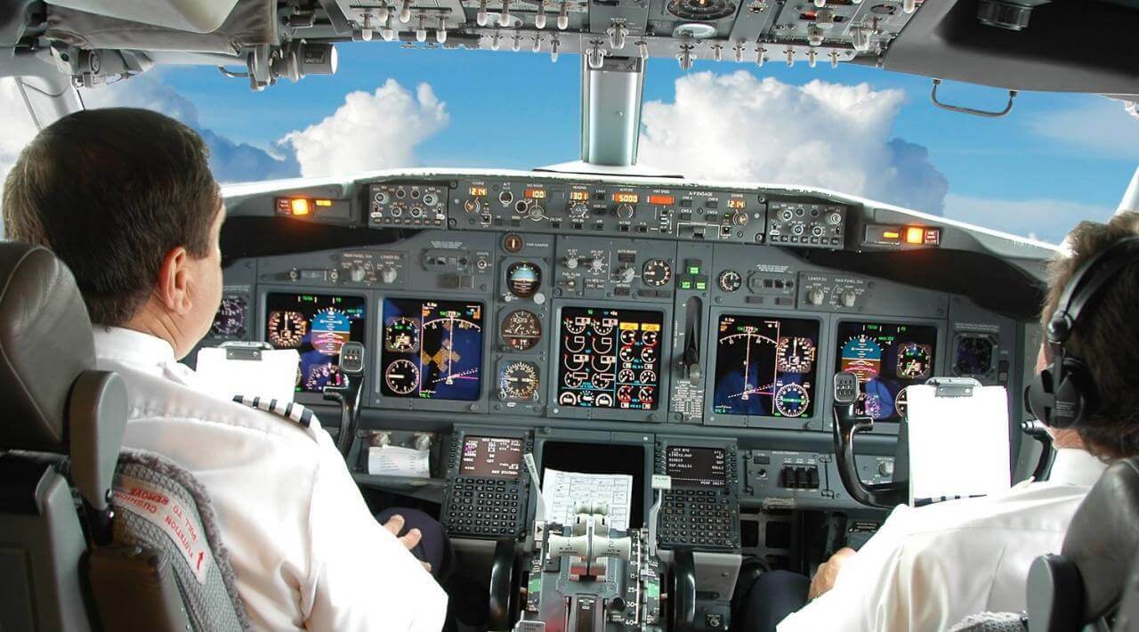 An over the shoulder image of two pilots flying an aircraft. You can clearly see the cockpit with the buttons and screens, as well as blue sky and clouds