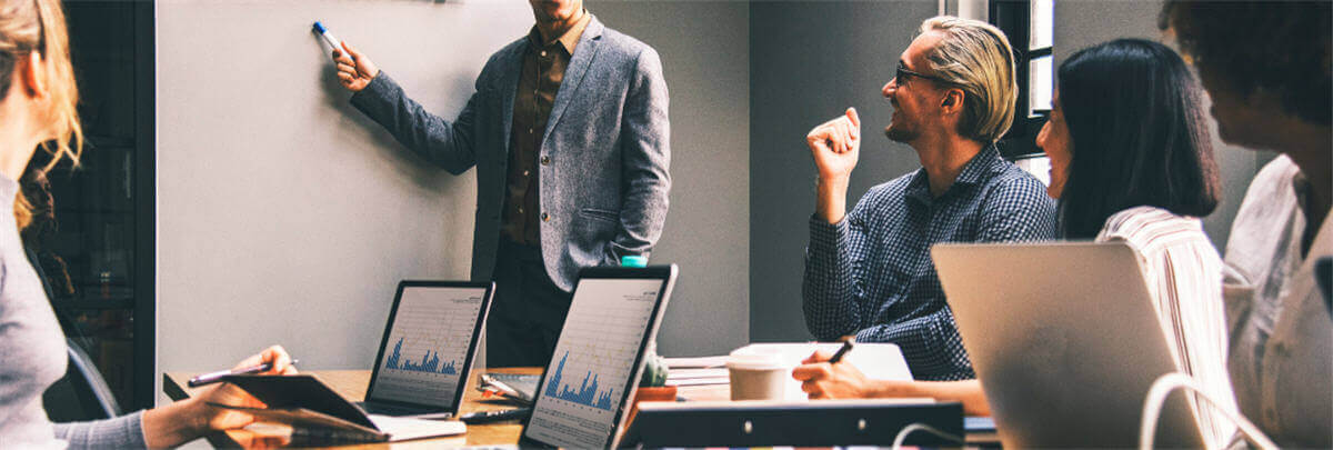 A group of smartly dressed professional sat around a table looking at someone pointing to a whiteboard. They are smiling and all sat with open laptops