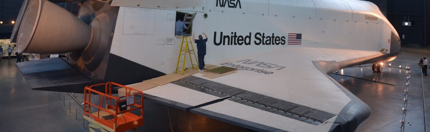 An image of a united states space rocket in a handgar. There is the USA flag on the side and two people working on the wing.